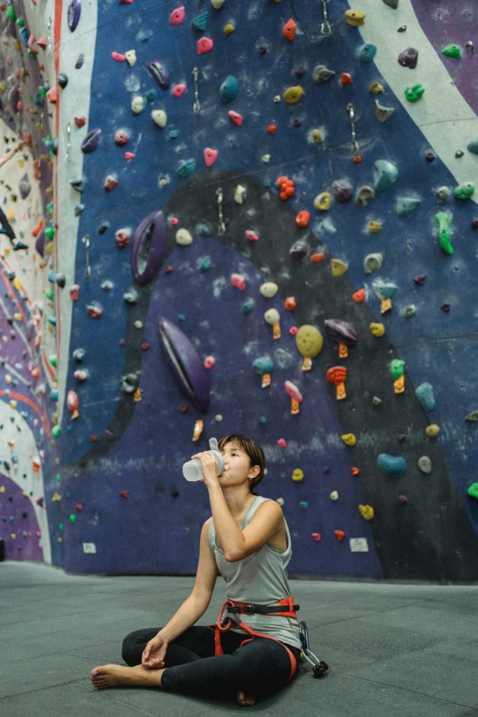 Women drinking from water bottle in front of climbing wall.